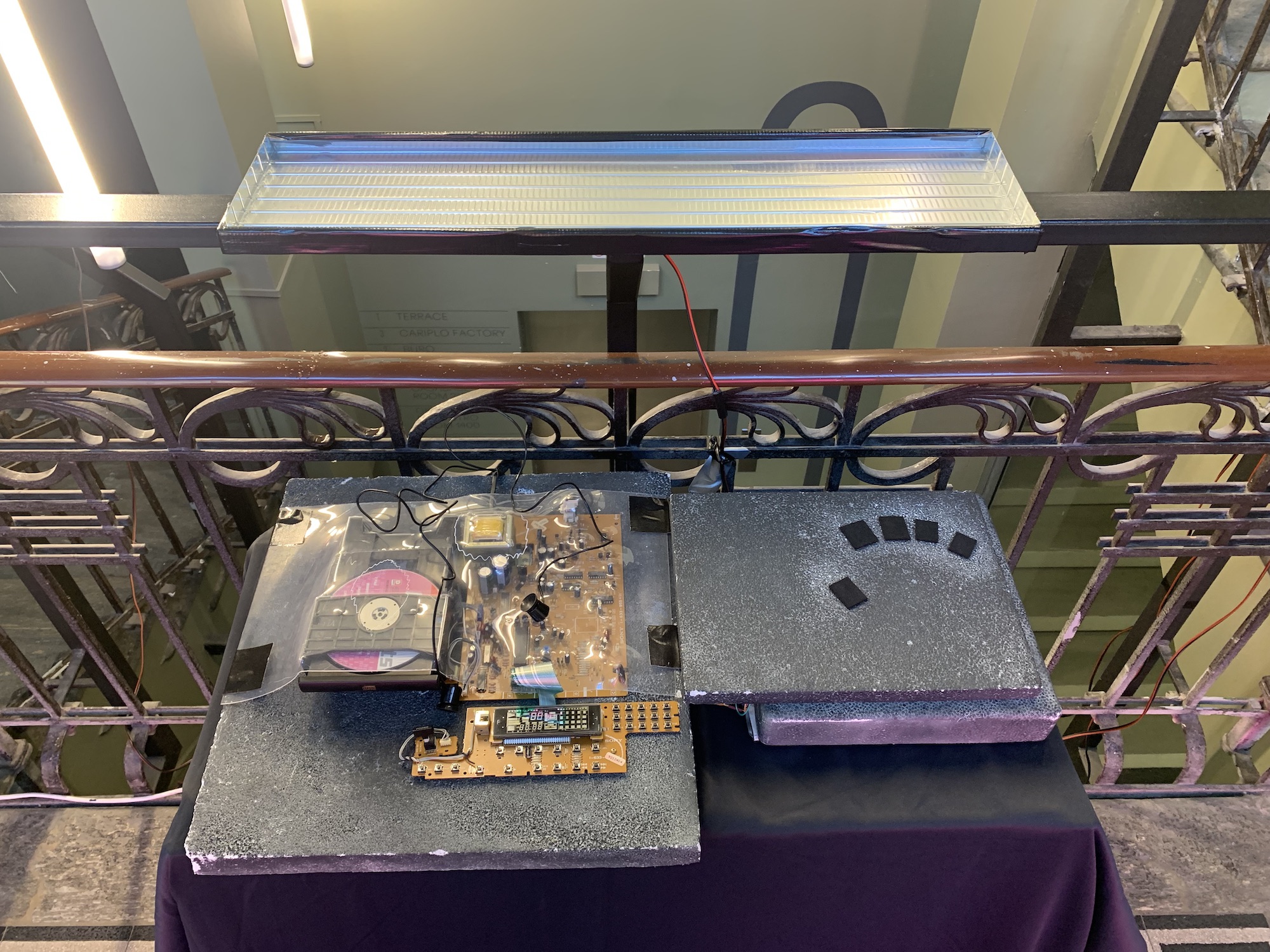 In a stairwell, a control surface and circuit boards on a table. Behind it on the railing is a shiny panel.